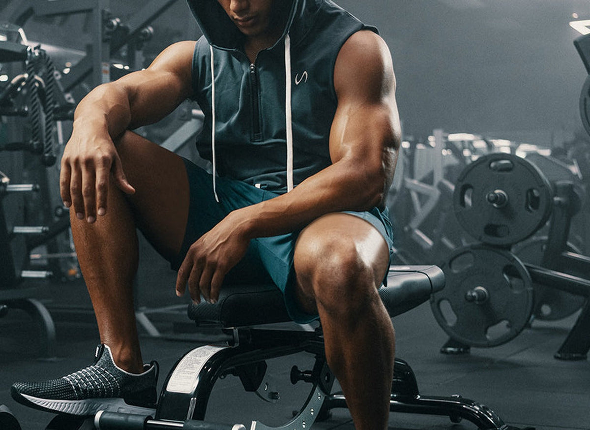Man in a sleeveless hoodie and teal shorts sitting on a gym bench