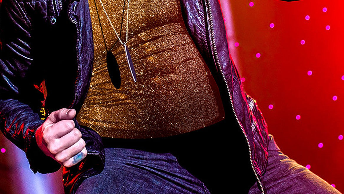 Man in a gold sequin top and leather jacket under red stage lights