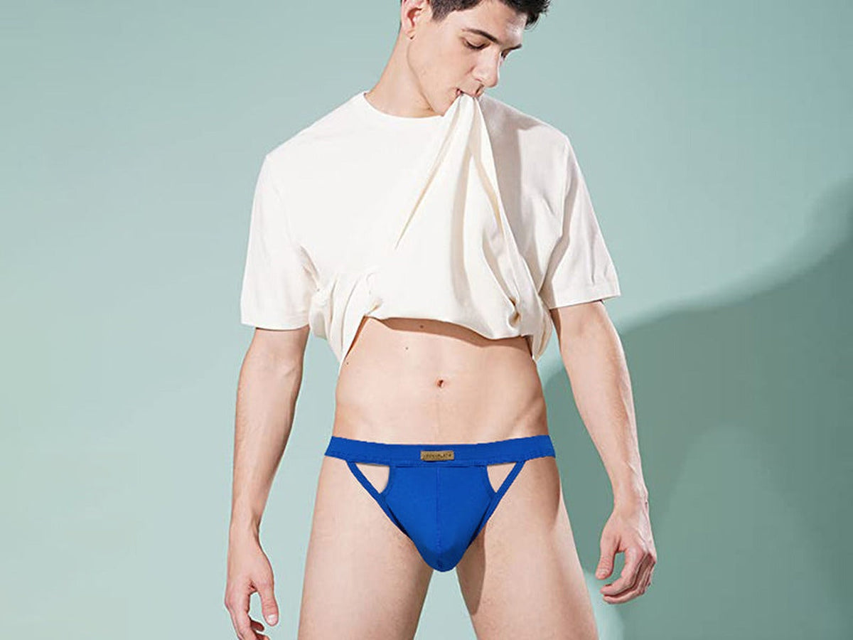 Man in a cream t-shirt wearing a blue Arjen Kroos jockstrap against a teal wall