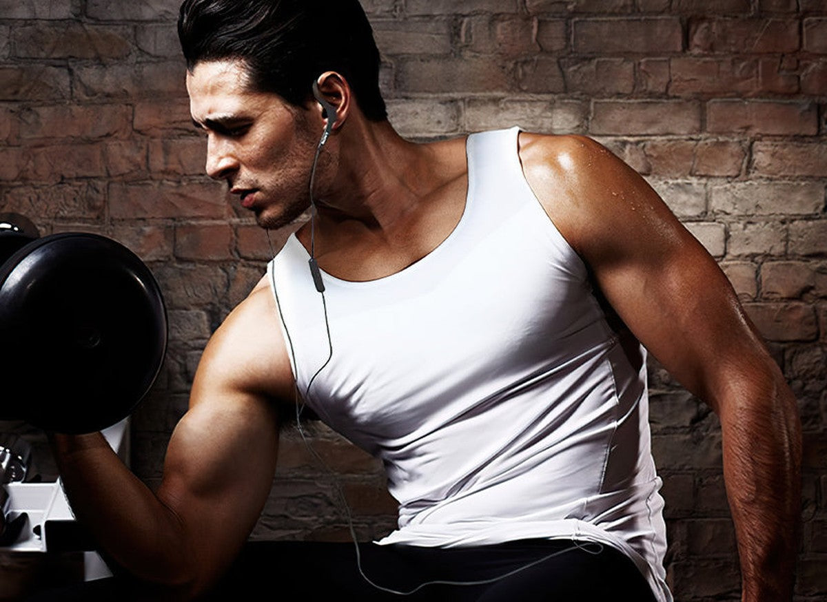 Man in a white compression tank top lifting a dumbbell in a brick-walled gym