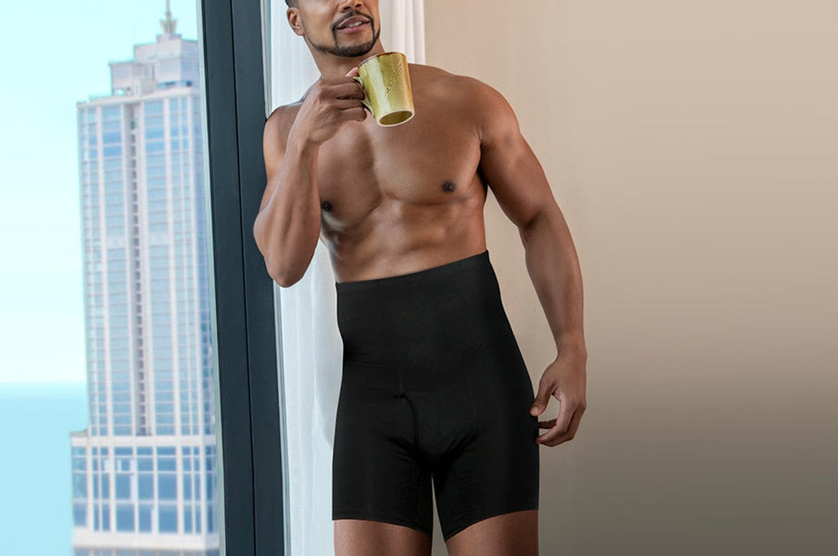 Man in black Arjen Kroos high-waist shapewear boxers holding a mug by a window with a city view