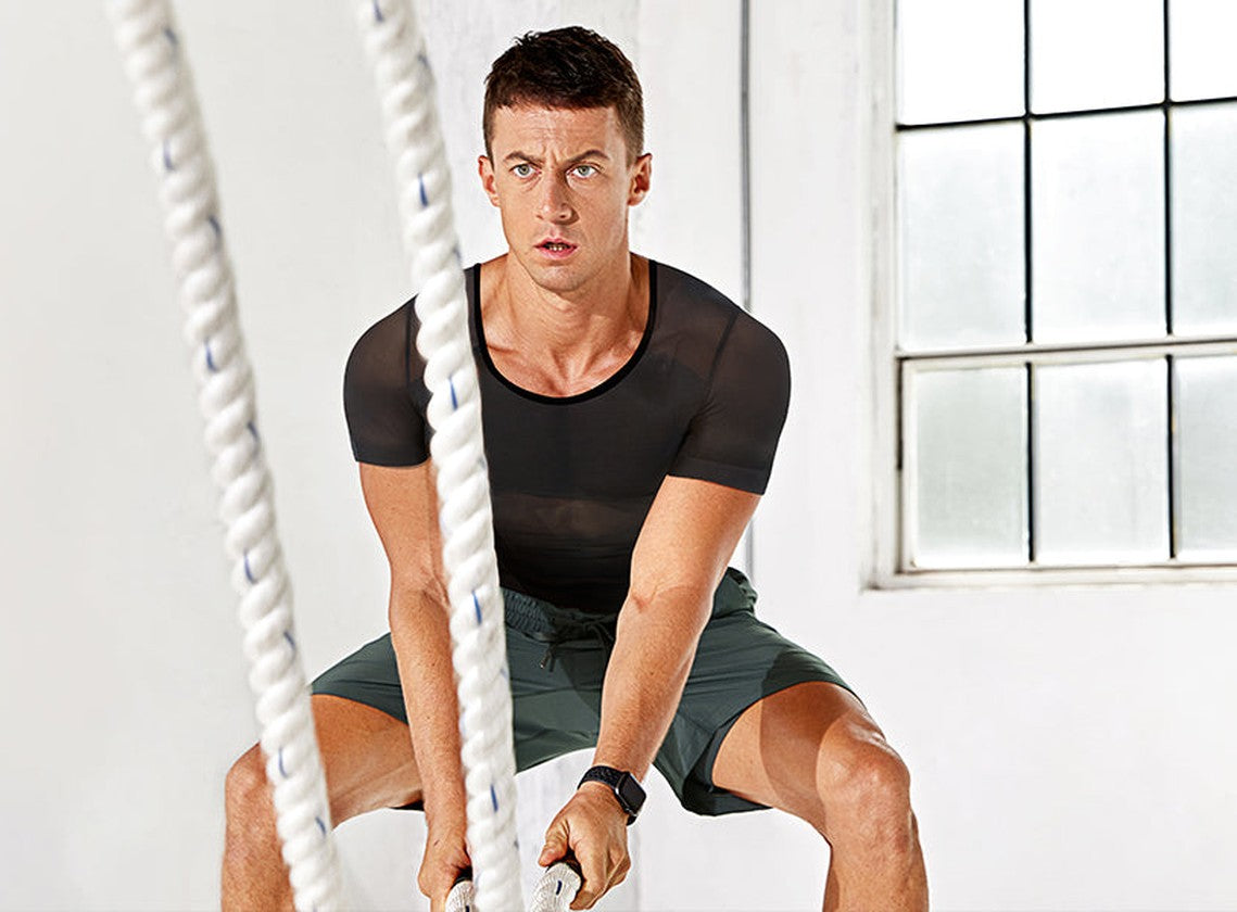 Man in a black short-sleeve compression undershirt training with battle ropes by a window